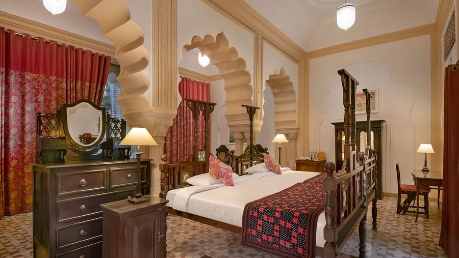A room with a four-poster bed, a dresser with a mirror, a chair, and arched doorways with decorative columns in Tijara Fort-Palace - 19th Century, Alwar.