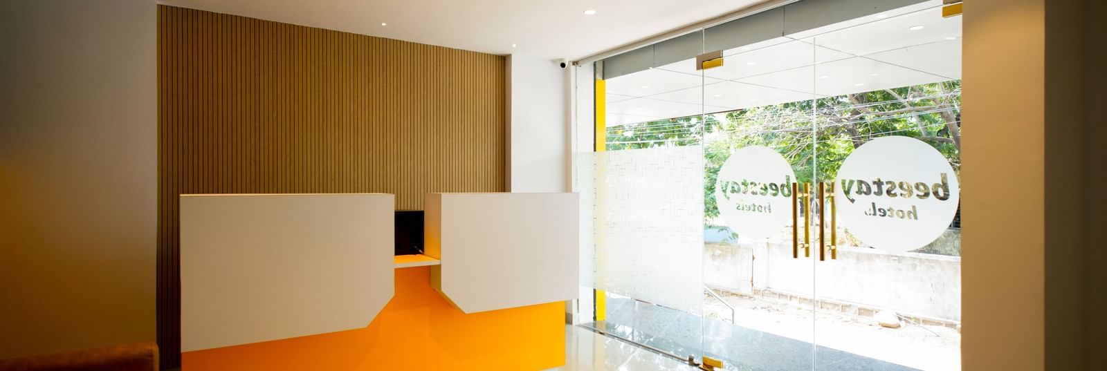 Side view of a modern hotel lobby with a bright orange and white reception desk, 2 brown armchairs, and a warm-toned wall behind them - Beestay Hotels Financial District, Hyderabad 