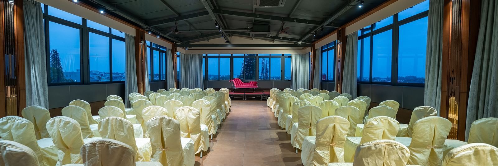 A view of the banquet hall at Hari Govinda's Express, Vijayanagar, with chairs arranged on either side of a central aisle leading to the stage, which features a cushioned sofa, while the curtains are drawn to the sides.