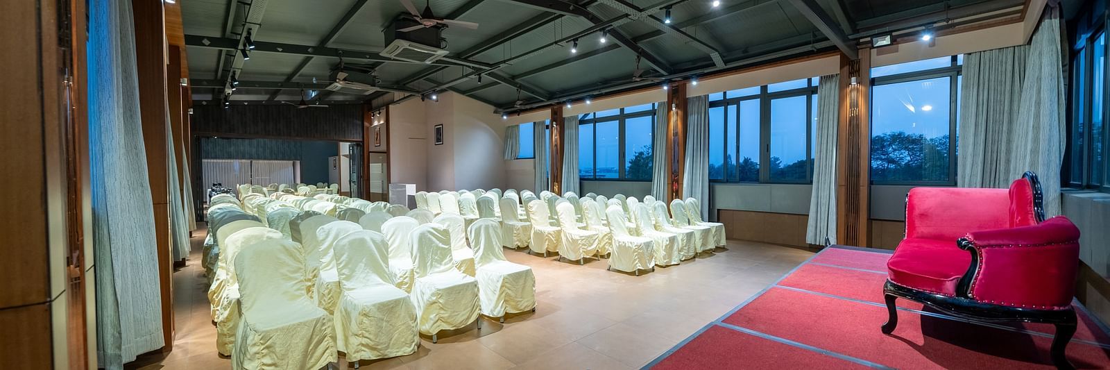 A view from near the stage at Hari Govinda's Express, Vijayanagar, reveals a central aisle with rows of chairs on either side, leading to a stage with a cushioned sofa.