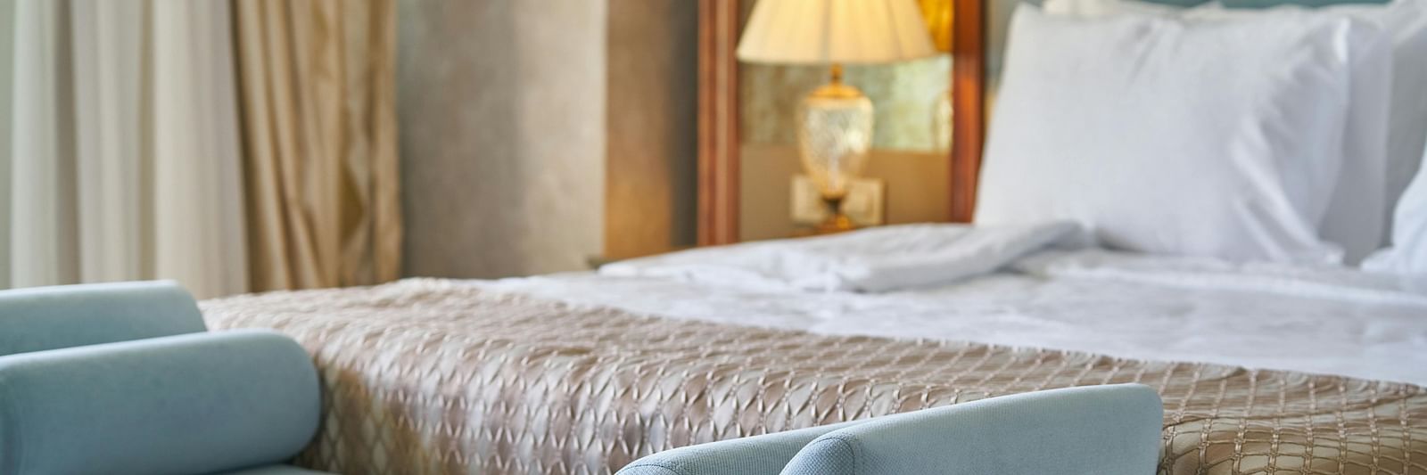 Elegant hotel bedroom featuring a plush light-blue bench at the foot of a well-made bed with a neutral quilt and a bedside lamp casting a warm glow.