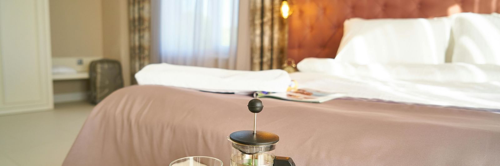 Well-lit hotel bedroom with king-size bed and a coffee tray featuring a French press, glass mug, and chocolate on a small table near the window.