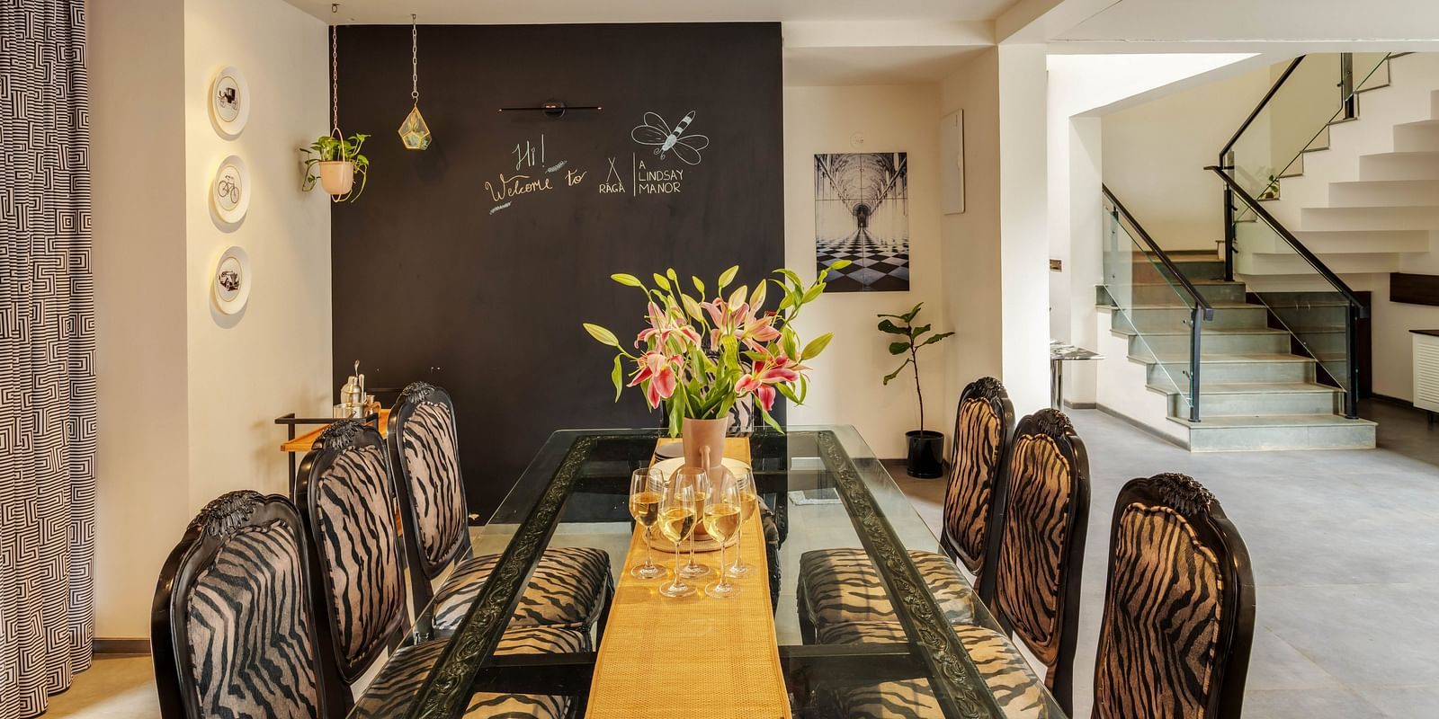  A dining setup at Lindsay Manor with a wooden-furnished table and chairs, a flower pot on the table, and a walkway leading to the staircase.