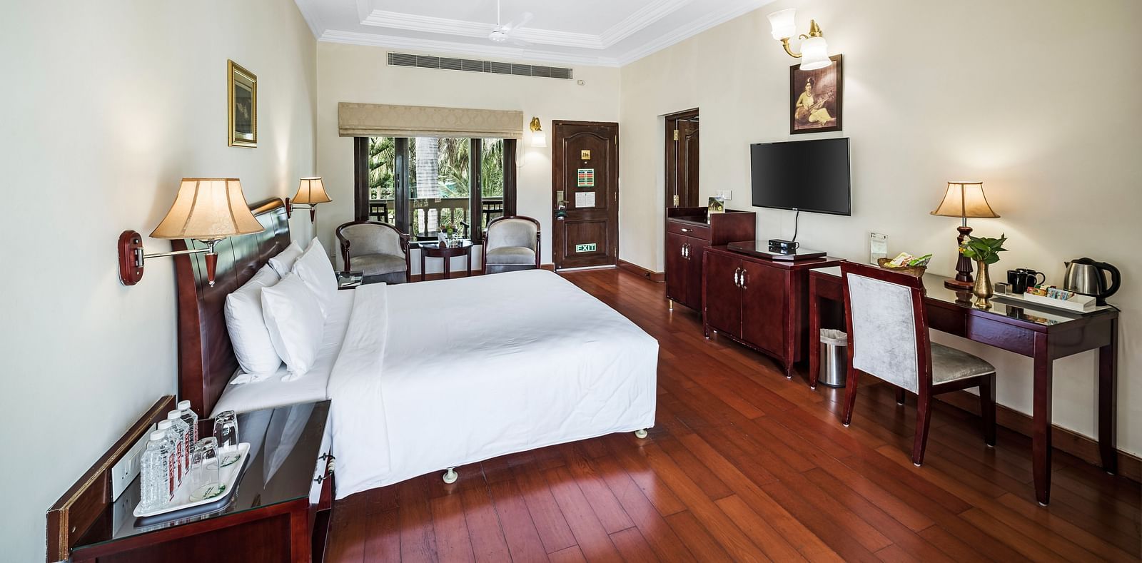 An image of a room with a king size bed, bed side table, lamps, flat screen TV, table, chairs, and modern amenities - GReaT Trails River View Resort Thanjavur By GRT Hotels