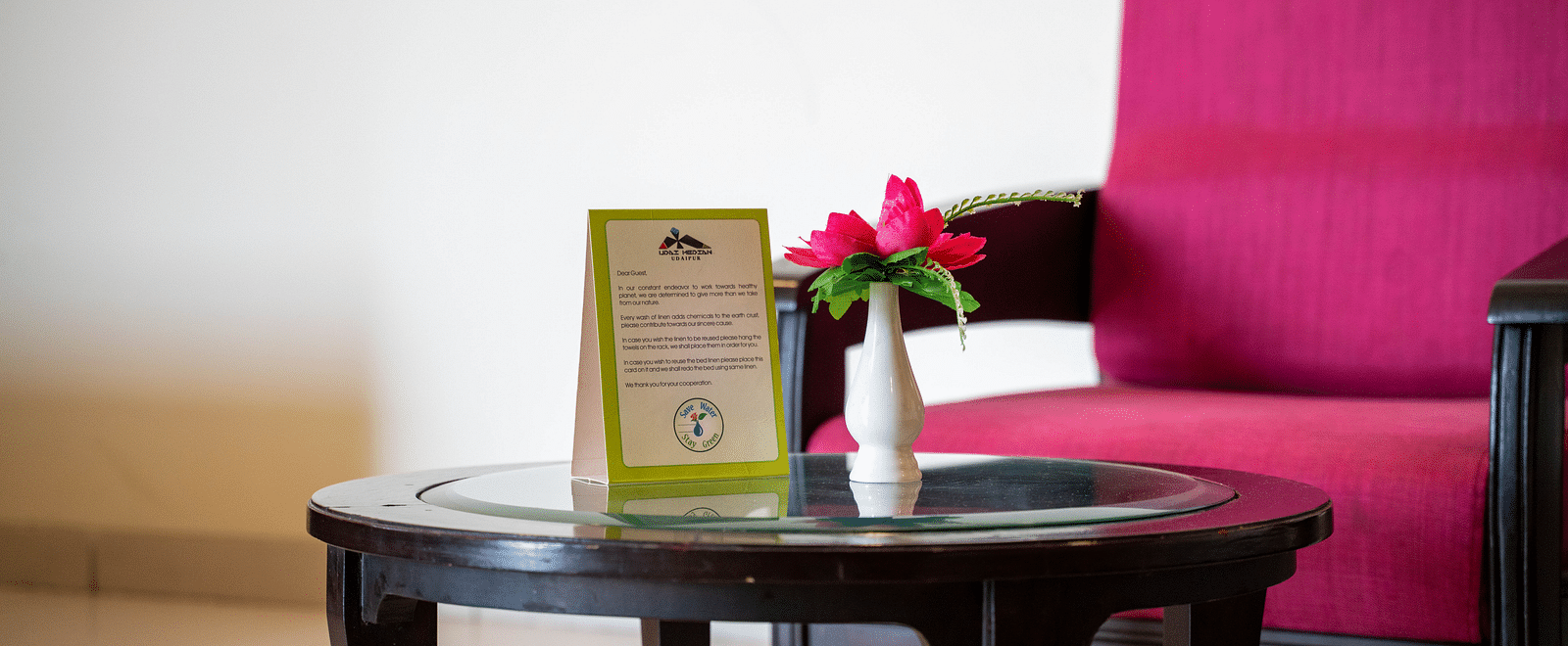 A small round table with a flower vase and a hotel service card, placed next to a armchair and a bed - Hotel Udai Median, Udaipur.