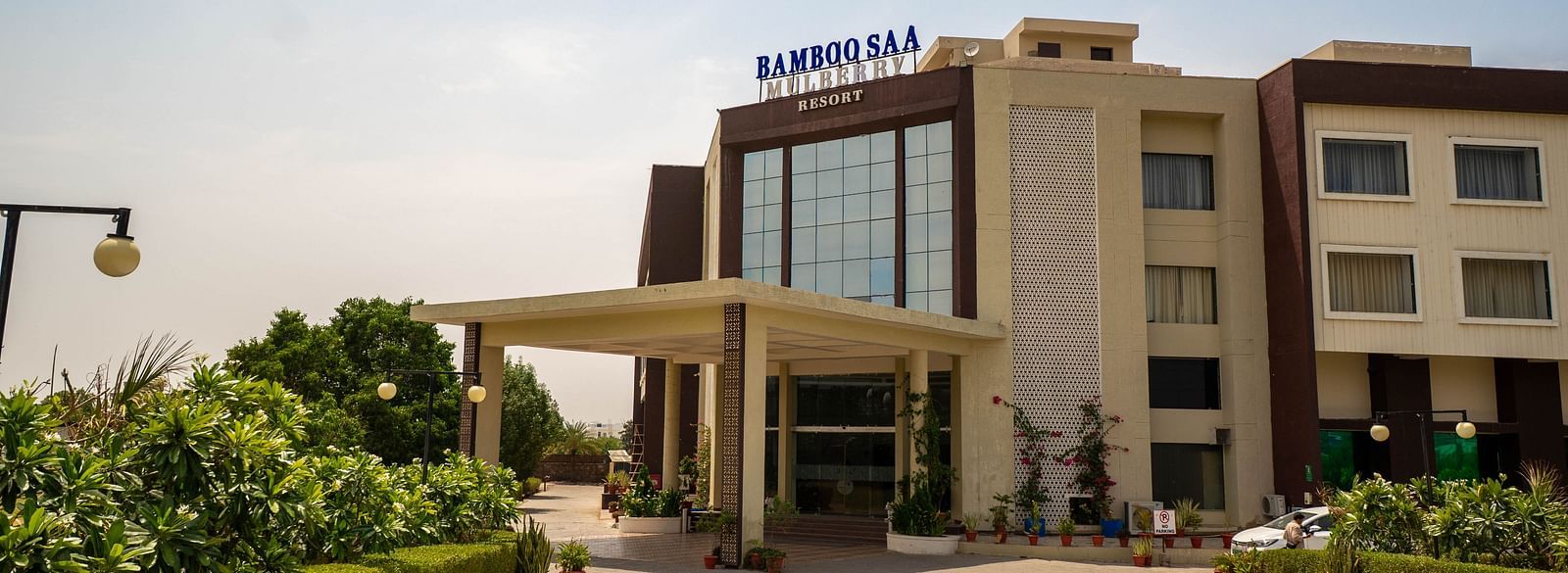 Driveway and extended Portico of Bamboo Saa Mulberry Resort in Pushkar surrounded by greenery on a sunny morning.