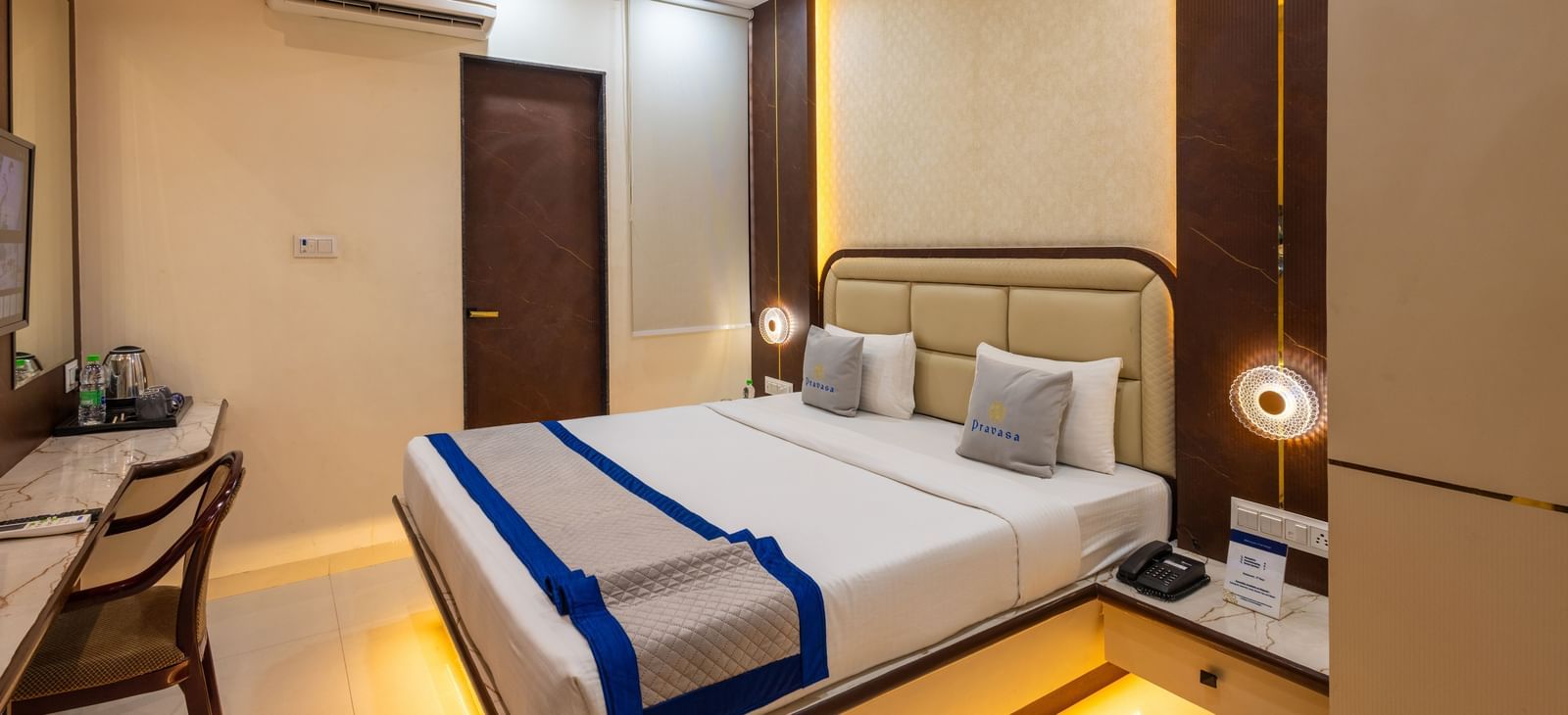 A room with a double bed, warm lighting, blue & beige runner, bedside lamps & a chair facing the wall at Pravasa Stay Dewas Naka, Indore.