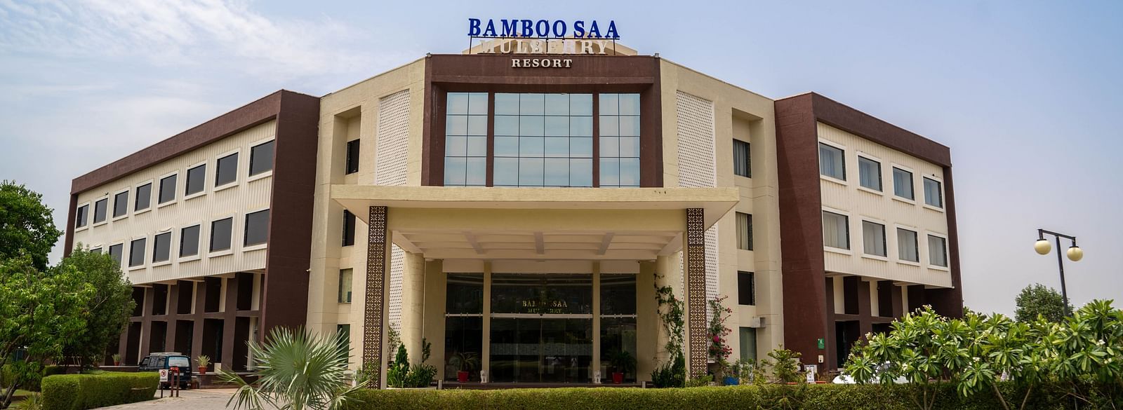 Front view of the grand facade and portico of Bamboo Saa Mulberry Resort, Pushkar with a large logo on a sunny day.