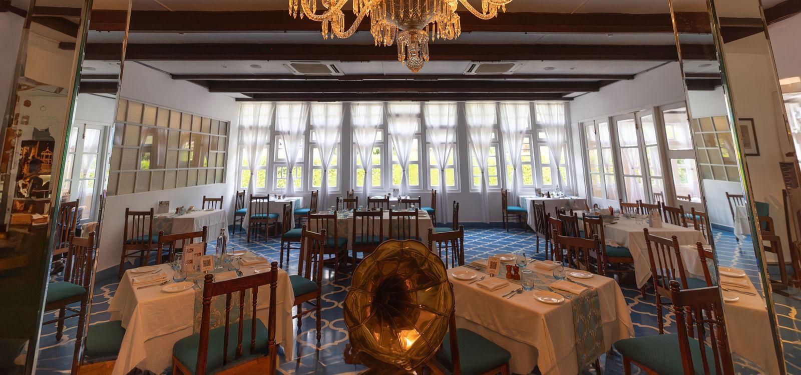 Dining arrangement in a restaurant at Saraca Hotel, Lucknow with large French windows in the backdrop and a chandelier hanging from the centre of the ceiling