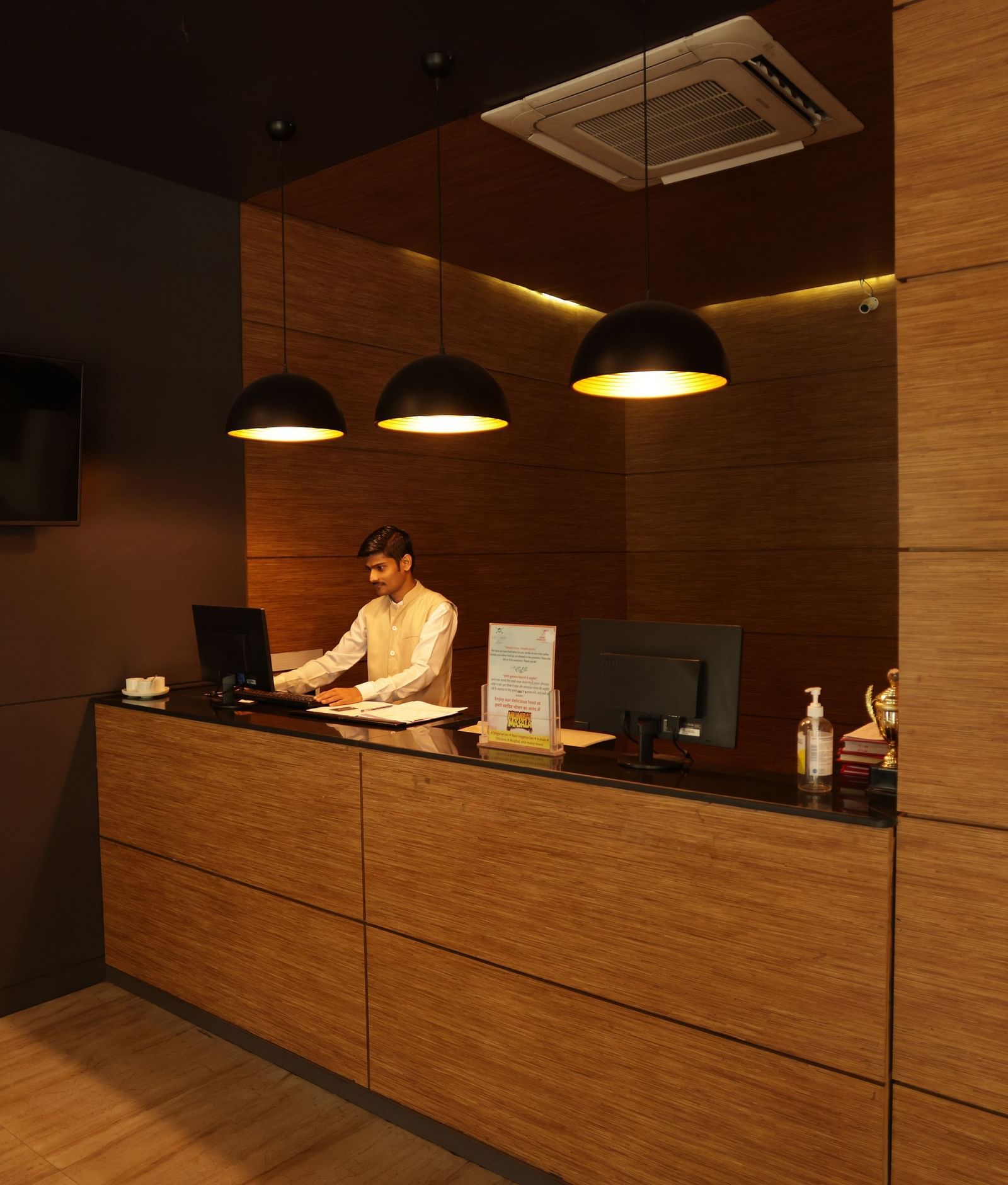 A person is front of the laptop at the front desk of the Max Hotel Jabalpur, featuring a wood-paneled reception area and warm lighting. 
