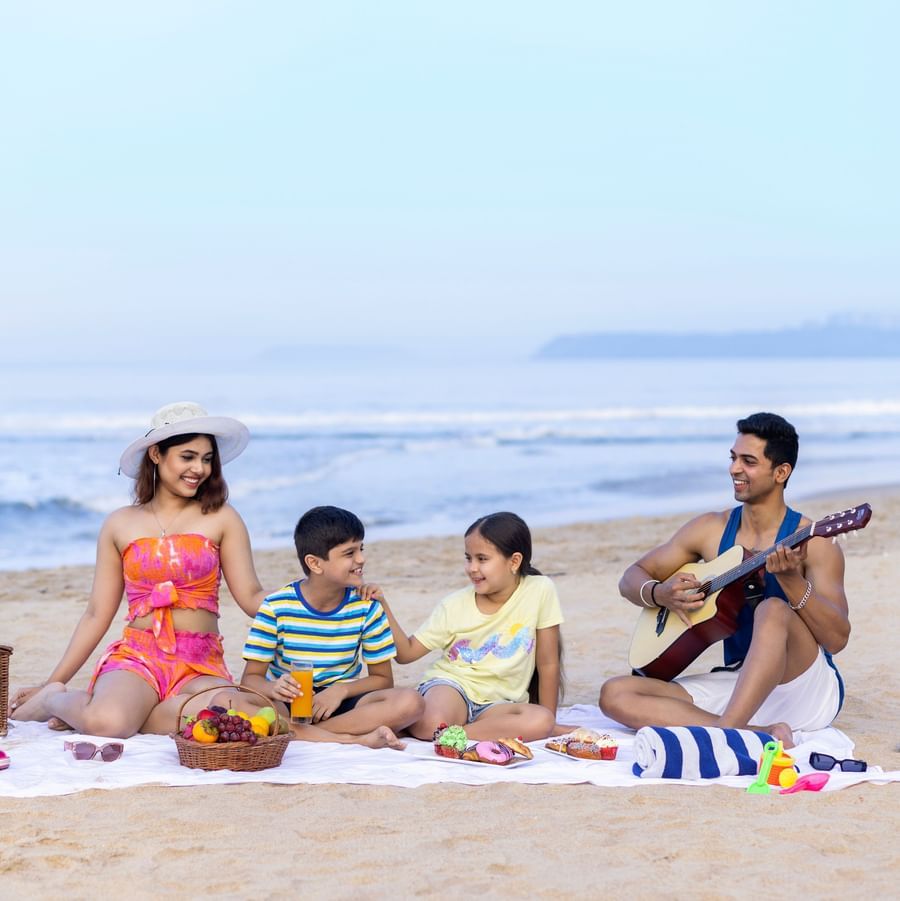 Two children enjoying with their parents on the beach side where a mat is placed and on top there are fruit baskets. The dad is playing a guitar.