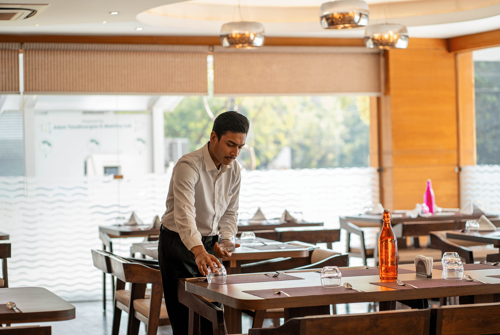 A staff member arranging tables in a restaurant with wooden interiors and large glass windows in the background - Hotel Udai Median, Udaipur.
