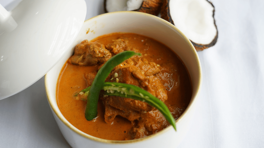 An overview of Chicken Xacuti served in a white claypot with coconut kept next to it.