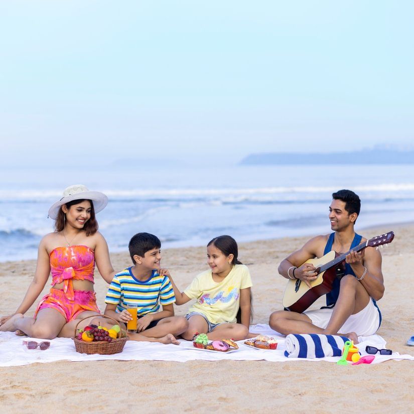 Two children enjoying with their parents on the beach side where a mat is placed and on top there are fruit baskets. The dad is playing a guitar.