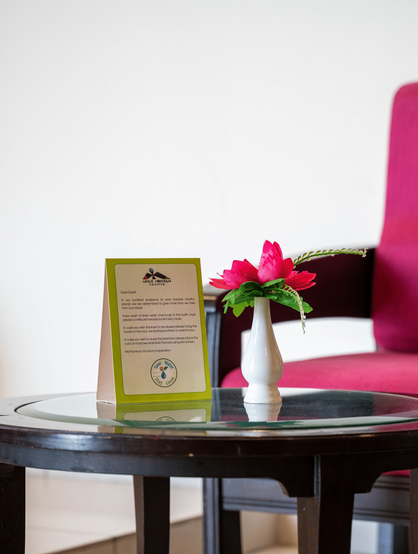 A small, round, dark wood table with a glass top holds a framed informational card and a small white vase with pink flowers, positioned in front of a bright pink armchair | Hotel Udai Median, Udaipur