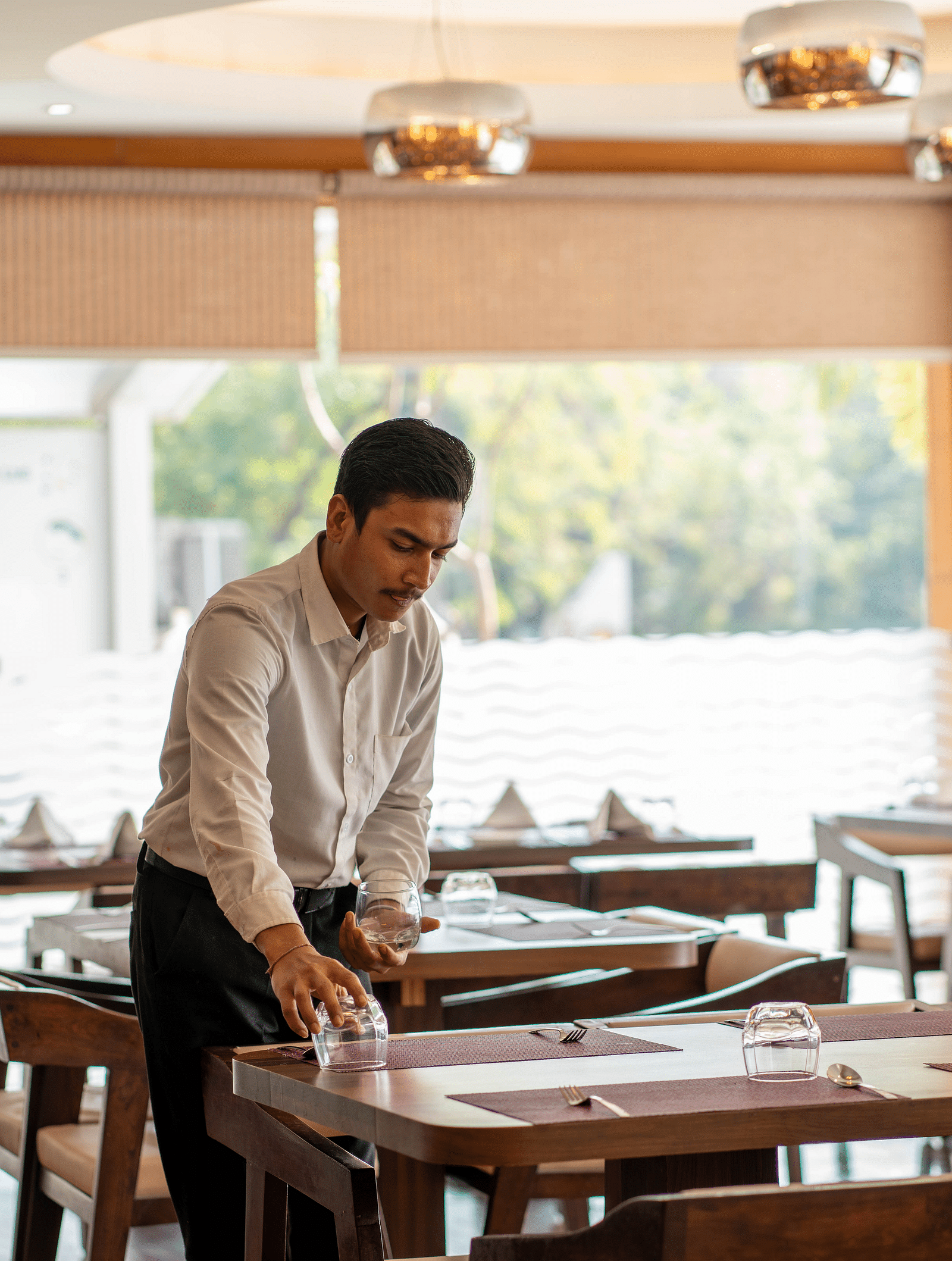 A waiter setting up a table in a restaurant with large glass windows | Hotel Udai Median, Udaipur