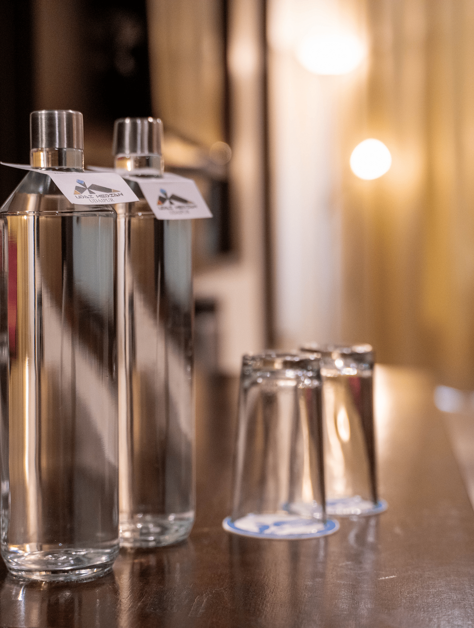 Two clear glass bottles with labels and two matching glasses filled with water are arranged on a polished surface, with a softly lit, warm-toned background | Hotel Udai Median, Udaipur