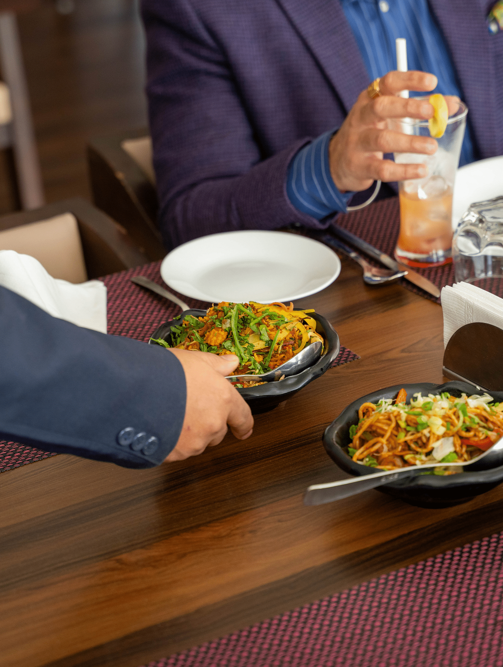 A person in a dark suit serving food from a bowl onto a table set with plates and glasses, where another person in a purple jacket is holding a drink | Hotel Udai Median, Udaipur