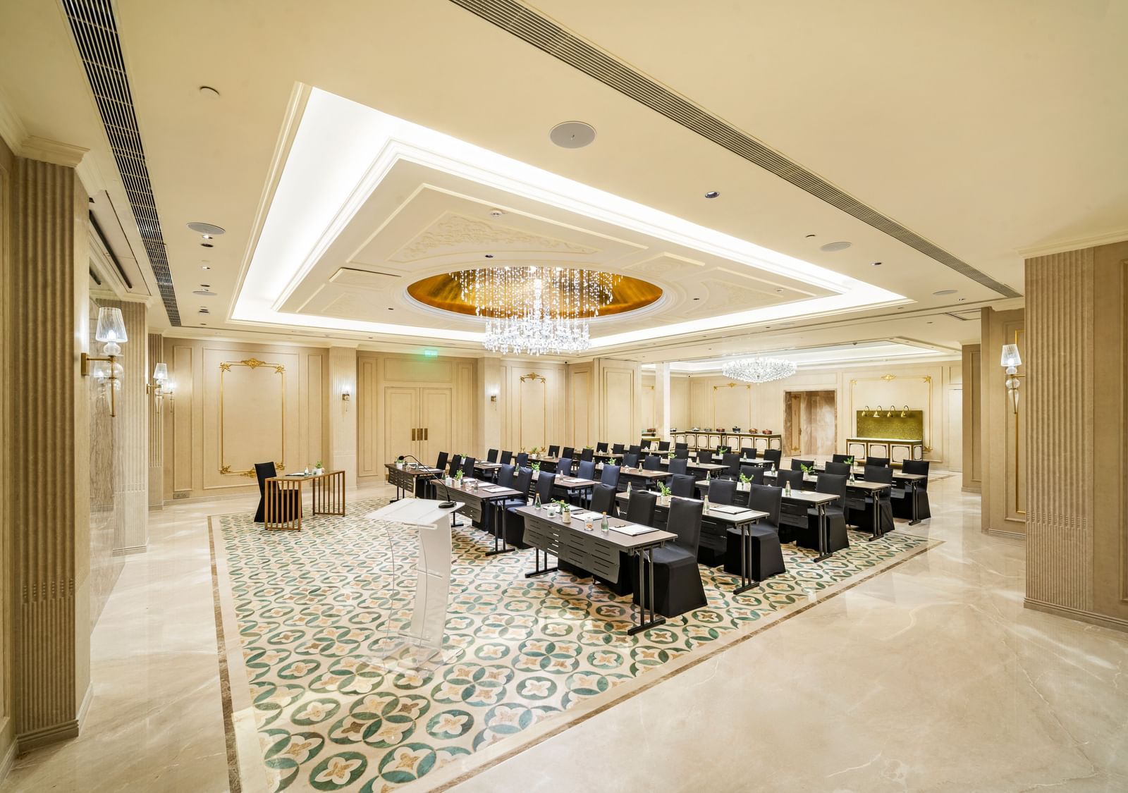A modern conference room set up in a classroom style at Grand Chennai showcasing multiple rows of dark tables and chairs facing forward. The geometric ceiling design and patterned carpet add contemporary flair to the professional space.