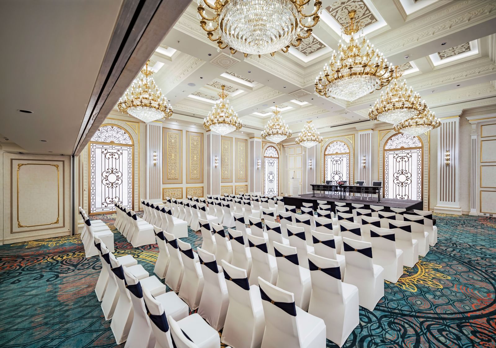 A grand hall prepared for a reception, showcasing elegant round tables with golden accents and white chairs, all beneath a ceiling adorned with multiple sparkling chandeliers.