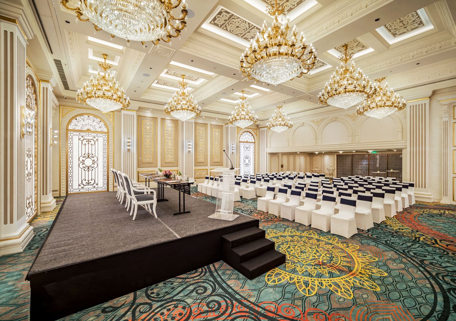A vibrant and dynamic event space, possibly a performance or fashion show venue, with a raised stage featuring unique furniture. The colorful patterned carpet and diverse lighting fixtures create an eclectic atmosphere.