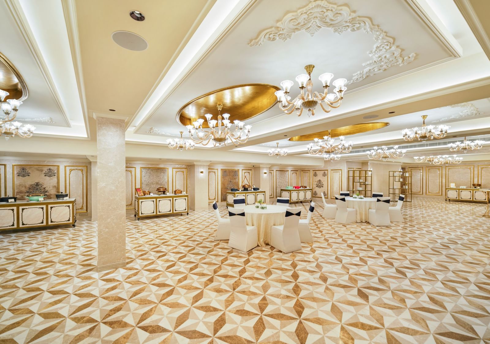 An elegant, spacious hall at Grand Chennai by GRT Hotels featuring a intricate tiled floor, high ceilings with decorative chandeliers, and several round cocktail tables, creating a sophisticated ambience perfect for a standing reception.