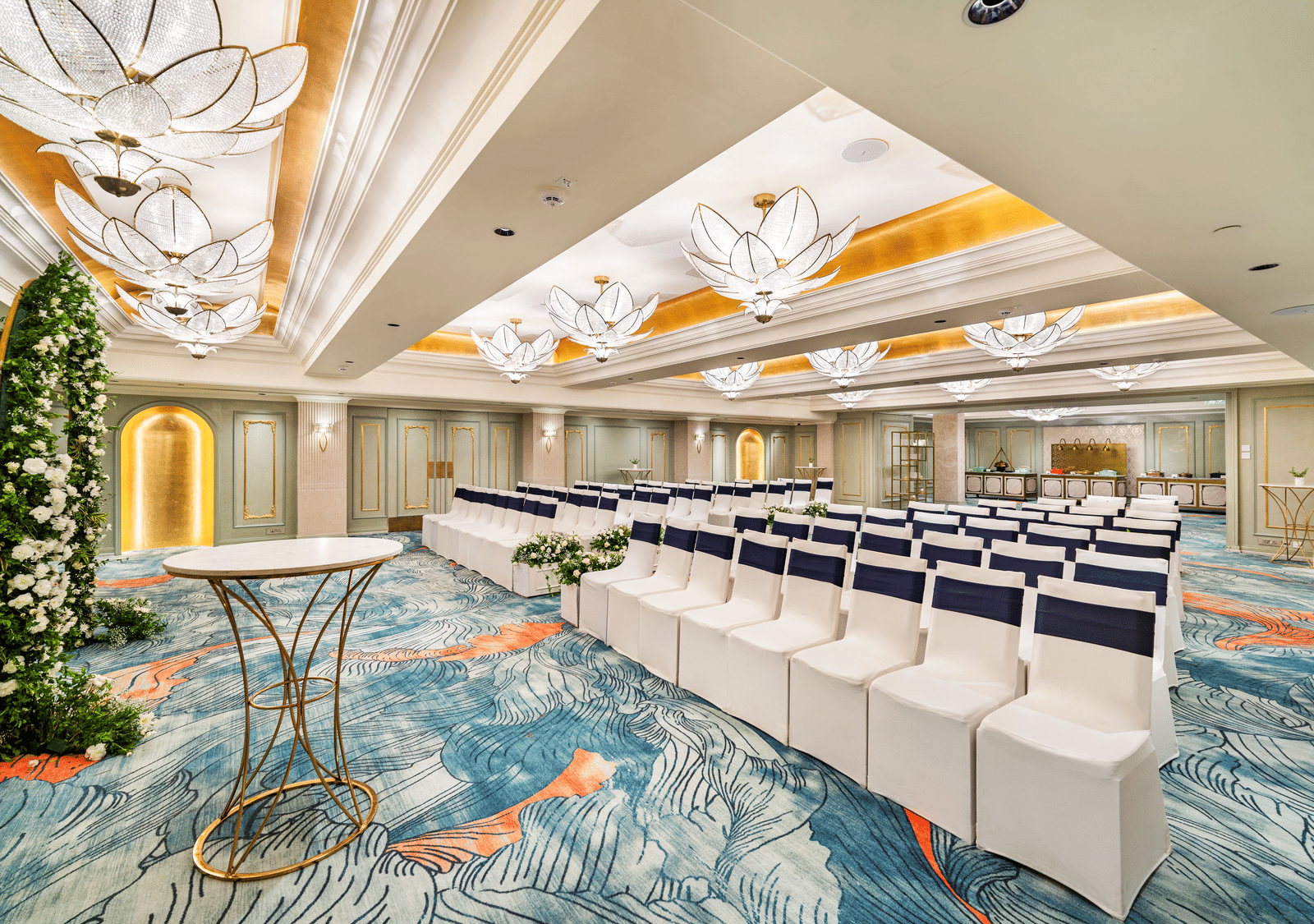 An expansive, well-lit ballroom at Grand Chennai by GRT Hotels configured for a seminar, featuring rows of pristine white chairs, a small stage area, and a striking tall plant as decor. The elegant ceiling and chandeliers add a touch of grandeur.