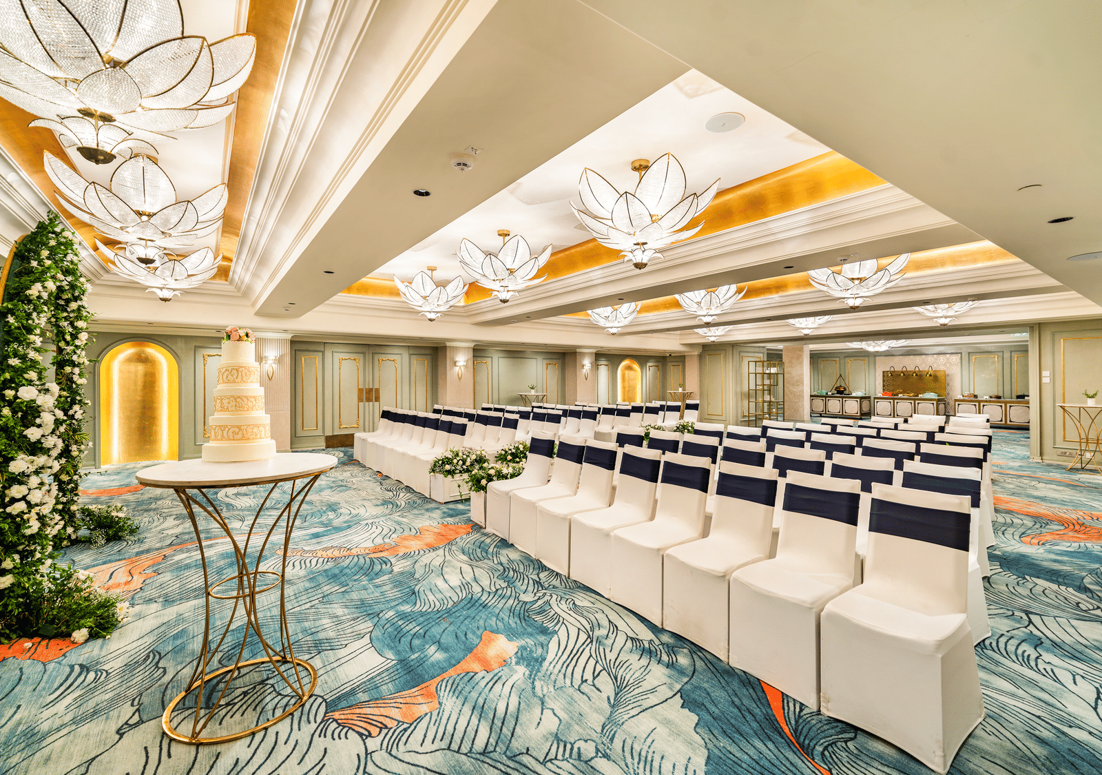 An event area at Grand Chennai by GRT Hotels, showcasing a tall cocktail table and neat rows of white chairs, all beneath the soft glow of exquisite chandeliers and an elegantly designed ceiling.