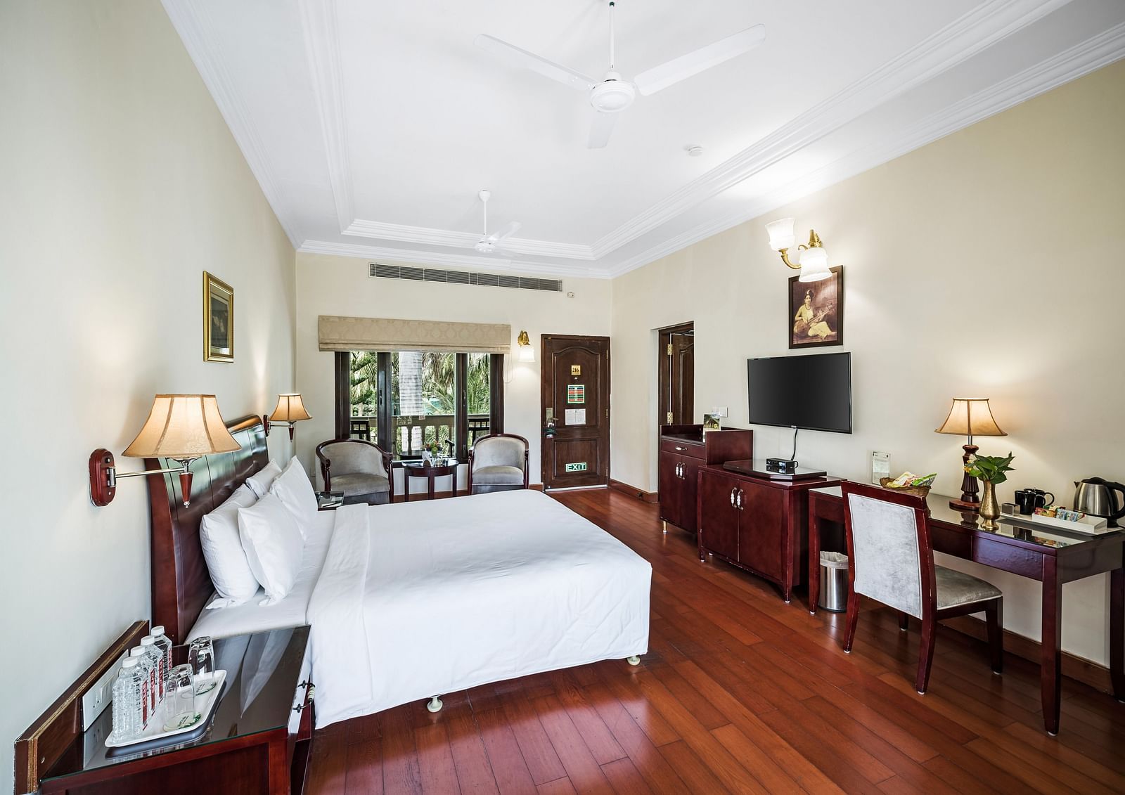 An image of a room with a king size bed, bed side table, lamps, flat screen TV, table, chairs, and modern amenities - GReaT Trails River View Resort Thanjavur By GRT Hotels