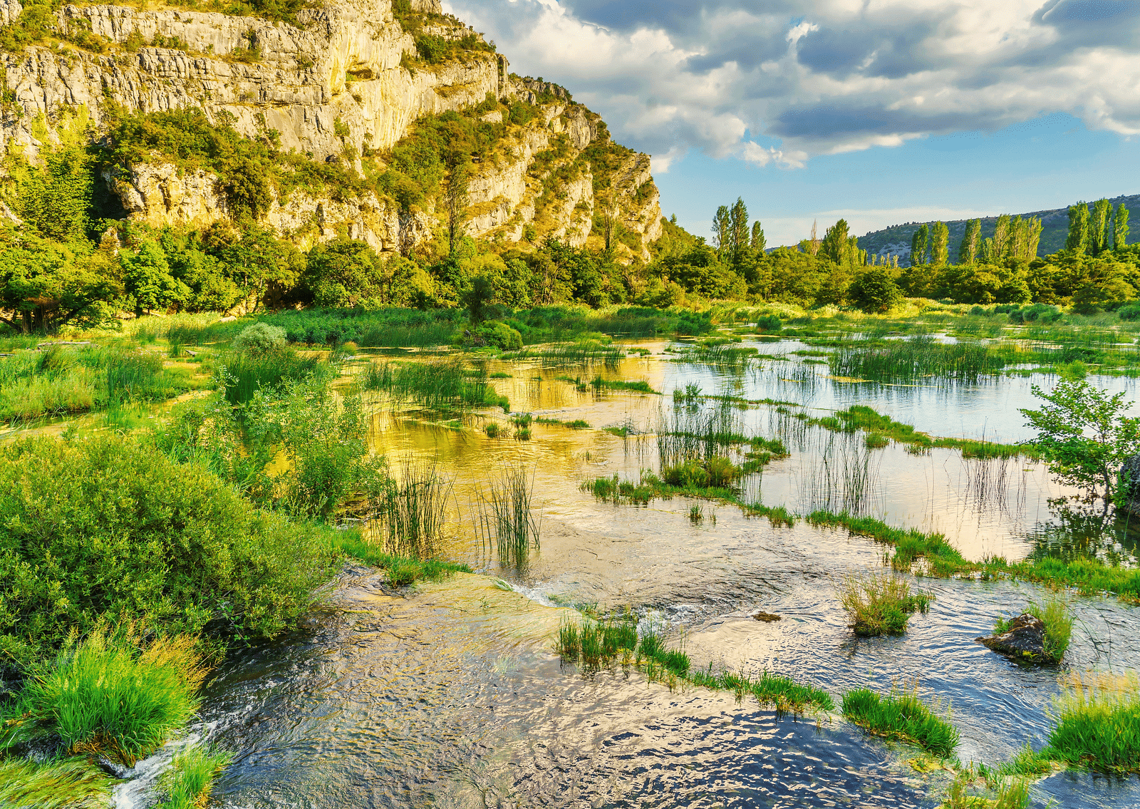 An overview of a small river with grass growing on natural steps and a hill in the background.