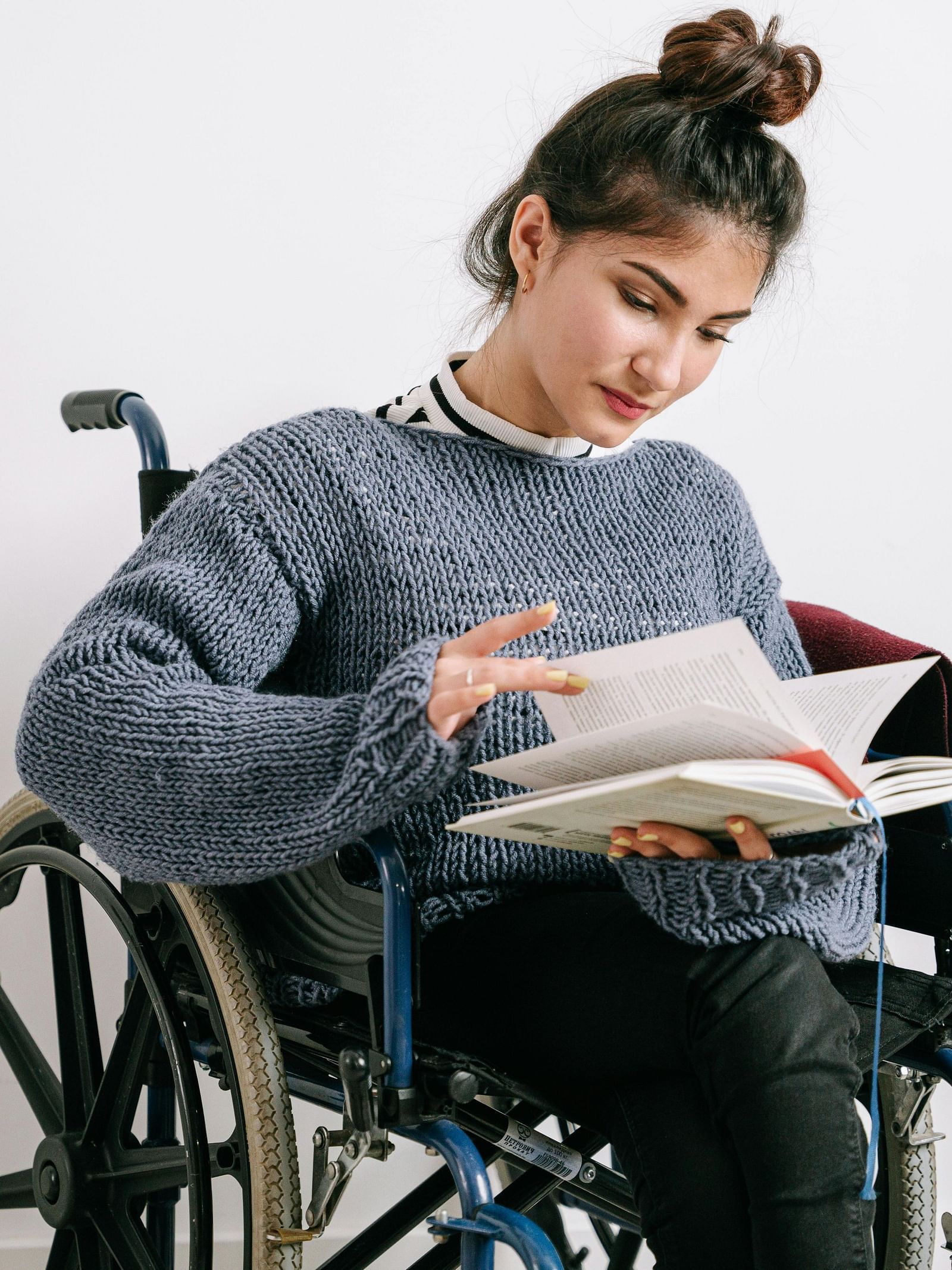 A young woman in a wheelchair reads a book intently against a plain white background.