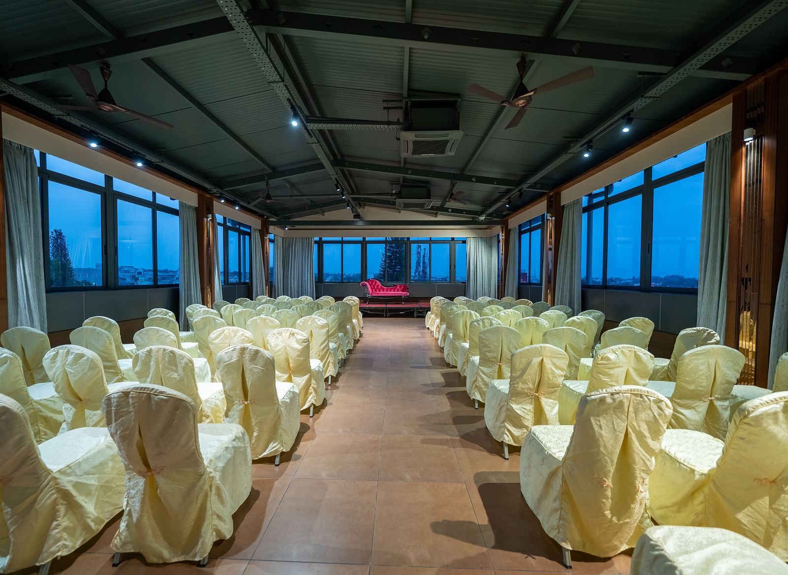 A view of the banquet hall at Hari Govinda's Express, Vijayanagar, with chairs arranged on either side of a central aisle leading to the stage, which features a cushioned sofa, while the curtains are drawn to the sides.