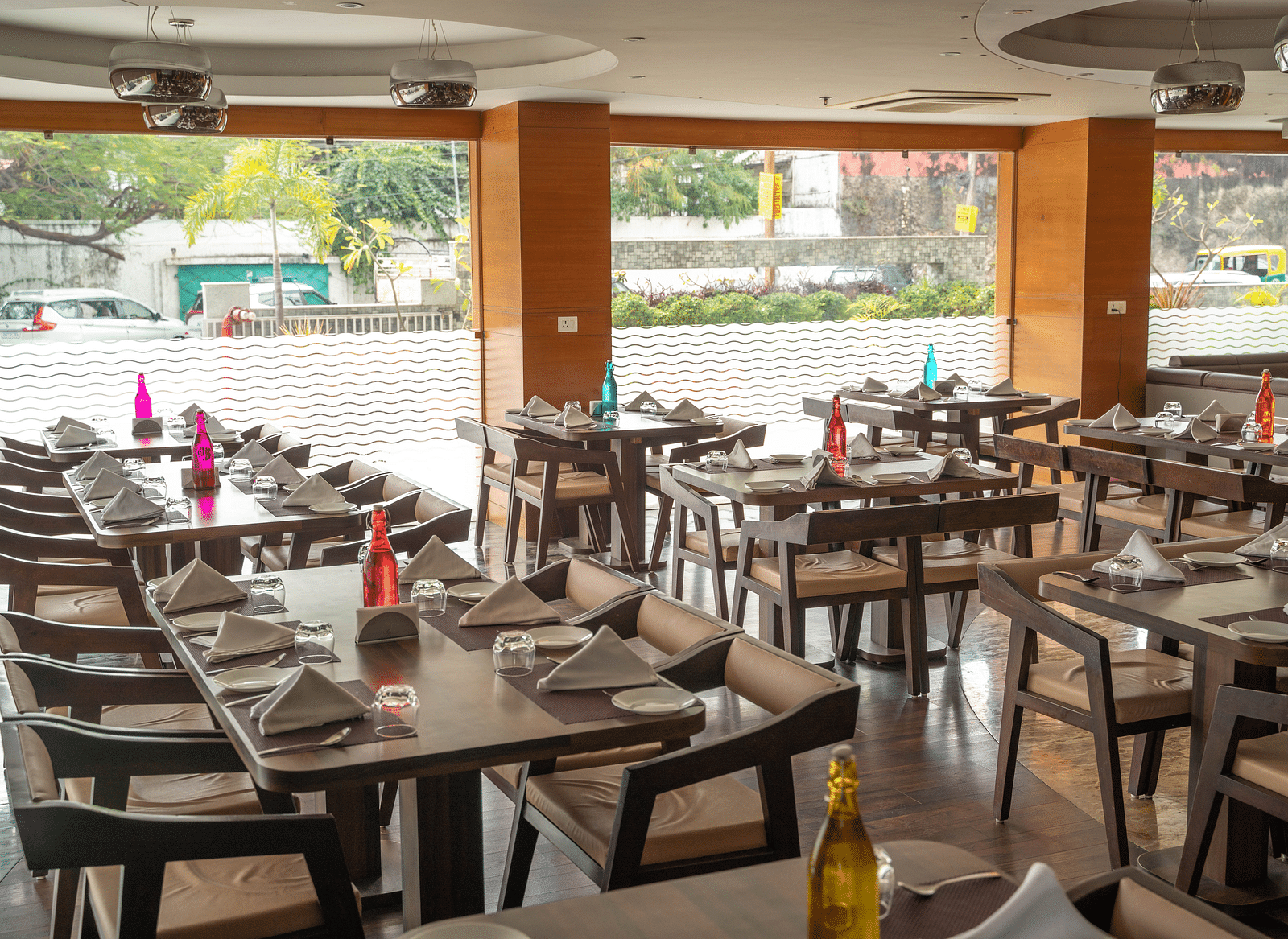 A bright dining area with wooden tables, comfortable seating, fresh flower arrangements and glass windows in the background - Hotel Udai Median, Udaipur.