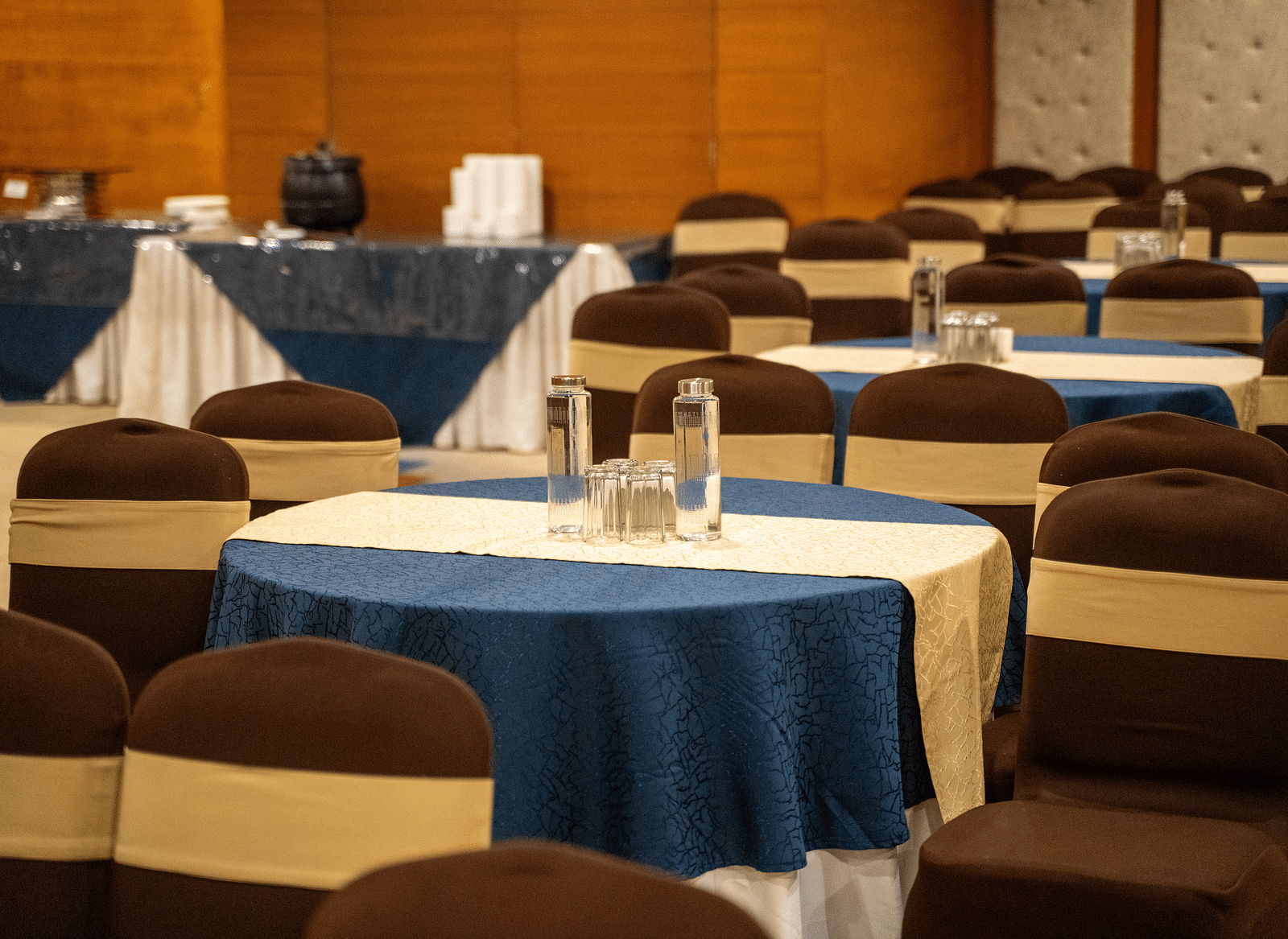 A banquet hall set up for an event, with neatly arranged round tables covered in blue tablecloths and brown chairs - Hotel Udai Median, Udaipur.
