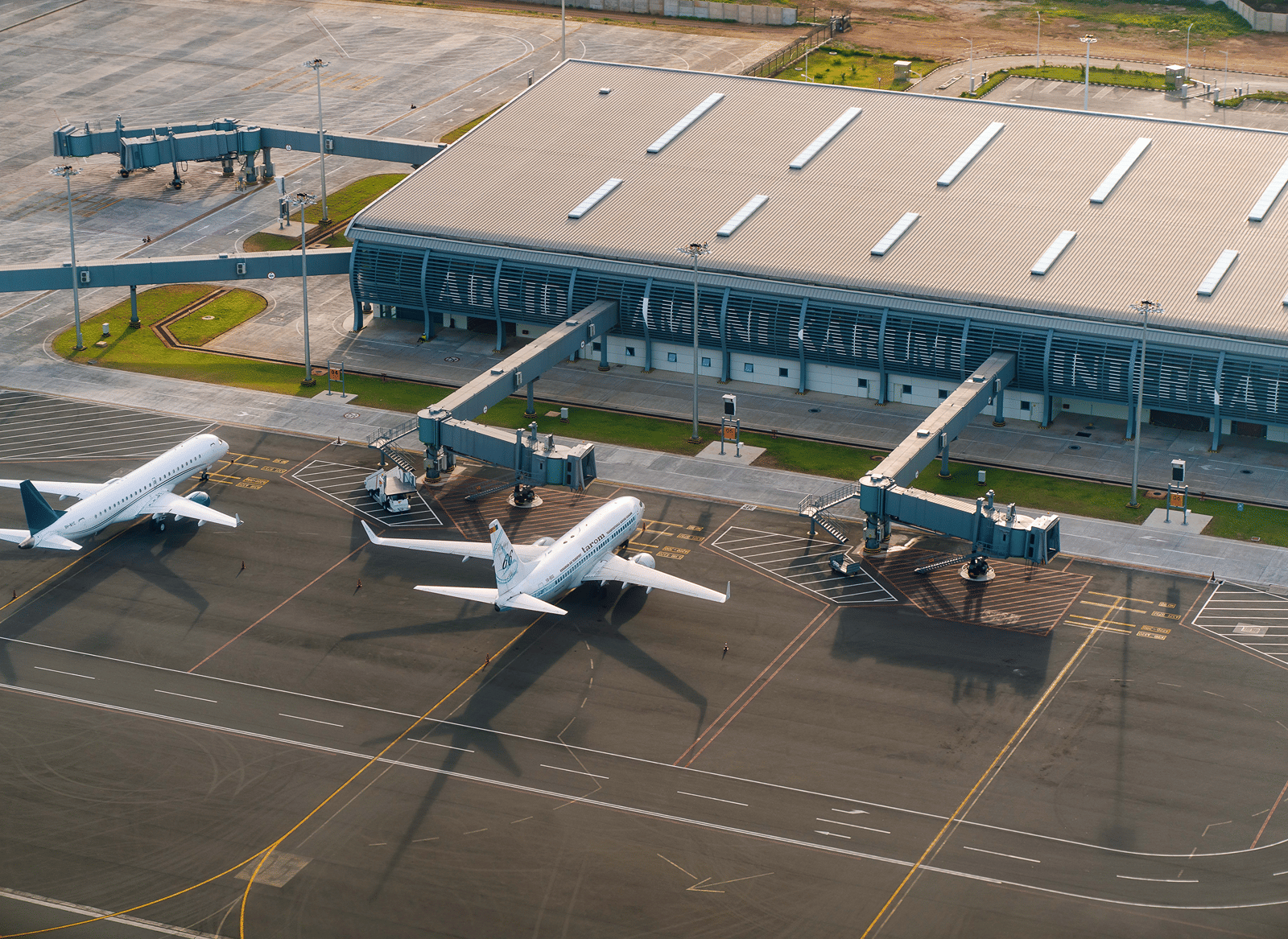 Aerial view of an airport terminal building with a corrugated roof, 3 jet bridges, and 2 white passenger aircraft parked on the apron in soft evening light.