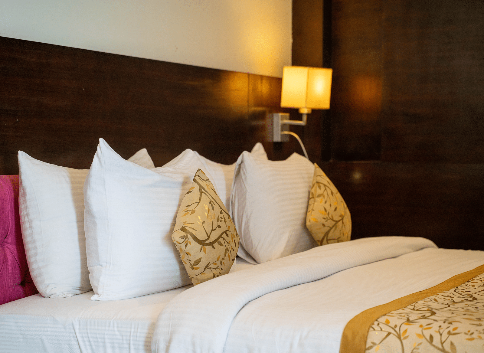 A neatly made bed with white linens and multiple pillows, including a patterned decorative pillow, is situated against a dark wood headboard, with a wall-mounted lamp emitting a warm light | Hotel Udai Median, Udaipur
