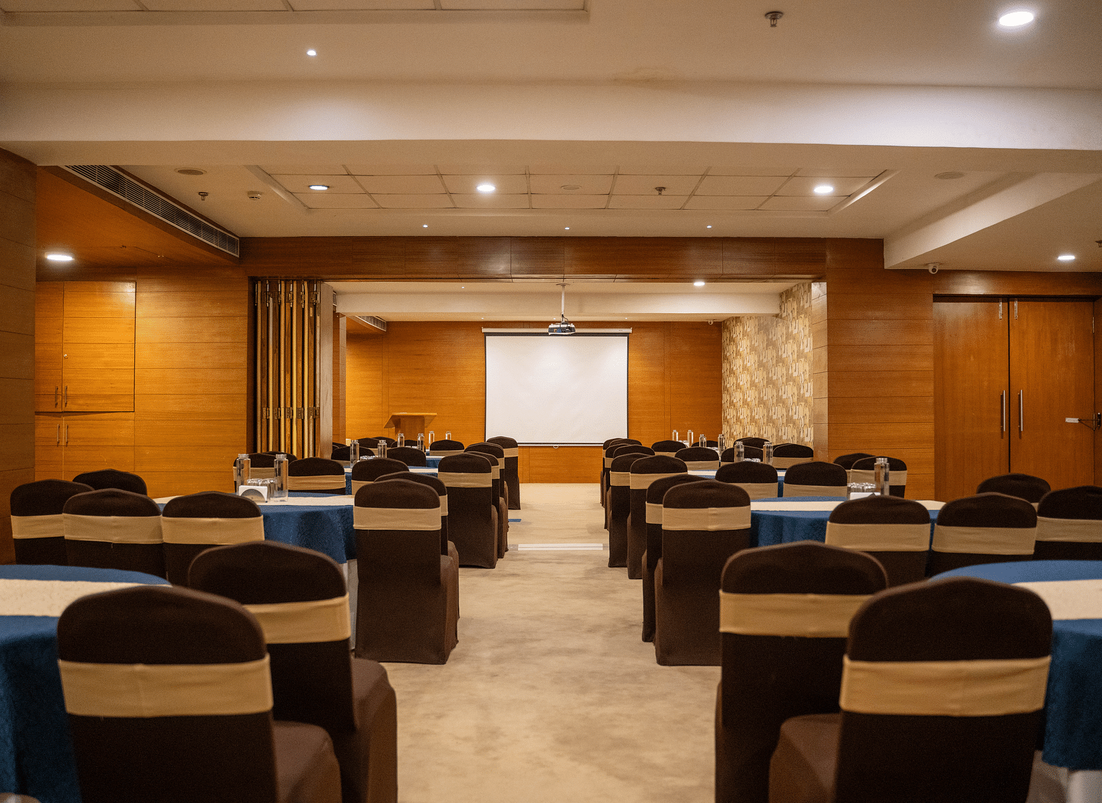 A well-lit, empty conference room features rows of dark brown chairs with light tan sashes, arranged around blue-clothed tables, facing a blank white projector screen at the room's far end | Hotel Udai Median, Udaipur