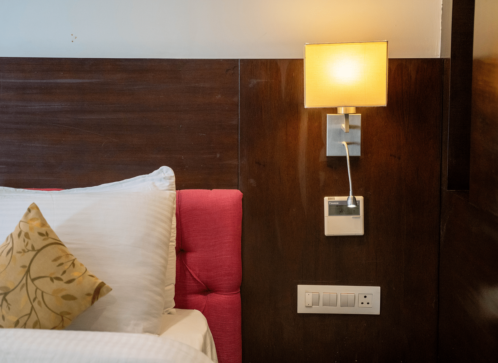 A close-up view showing a portion of a hotel room, featuring a white pillow with a decorative patterned cushion resting against a red headboard, beside a wall-mounted lamp with a warm, glowing light | Hotel Udai Median, Udaipur