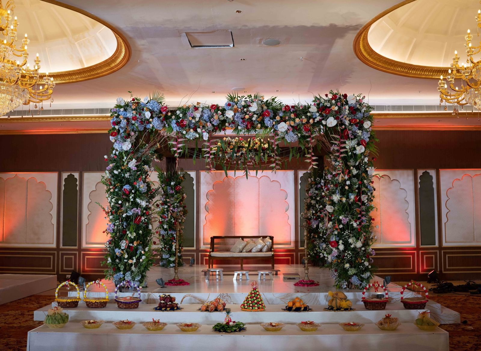 A stage with couch, an arch with ornamental flowers & pants and chandeliers hanging from the ceiling - Kaldan Samudhra Palace