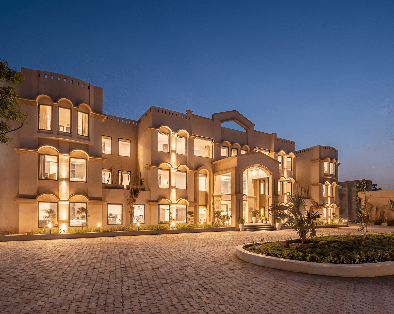 An image of a well lit facade with a huge concrete lawn and a few trees - Brij Eternity, Vrindavan