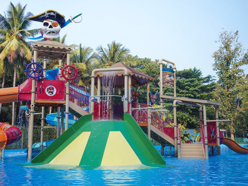 A colourful pirate-themed water playground with slides, splash features, and a skull-shaped structure at a water park, surrounded by palm trees.