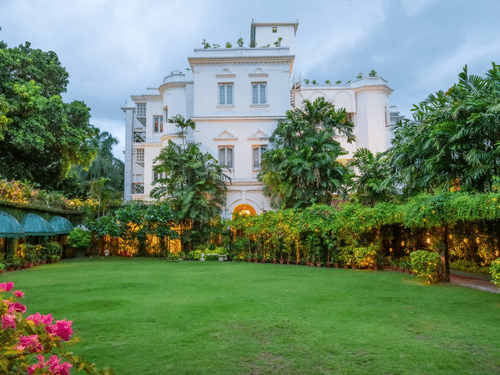 Facade view of Kenilworth Hotel, a city centre hotel in Kolkata, with a lawn in front of it, trees having fairy lights, many trees surrounding the lawn area as seen during twilight hour.