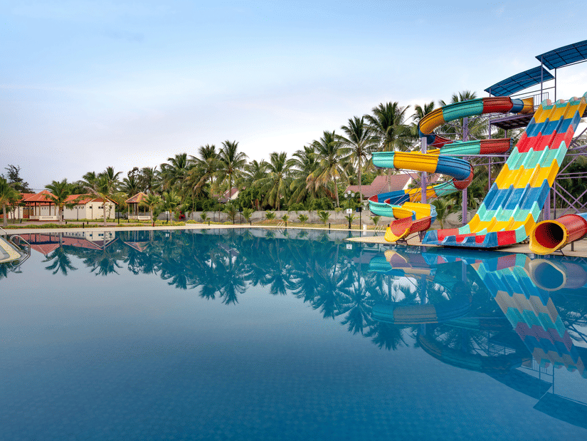 A view from a waterbody of a water park with circular and straight slides on the right hand side and trees in the background