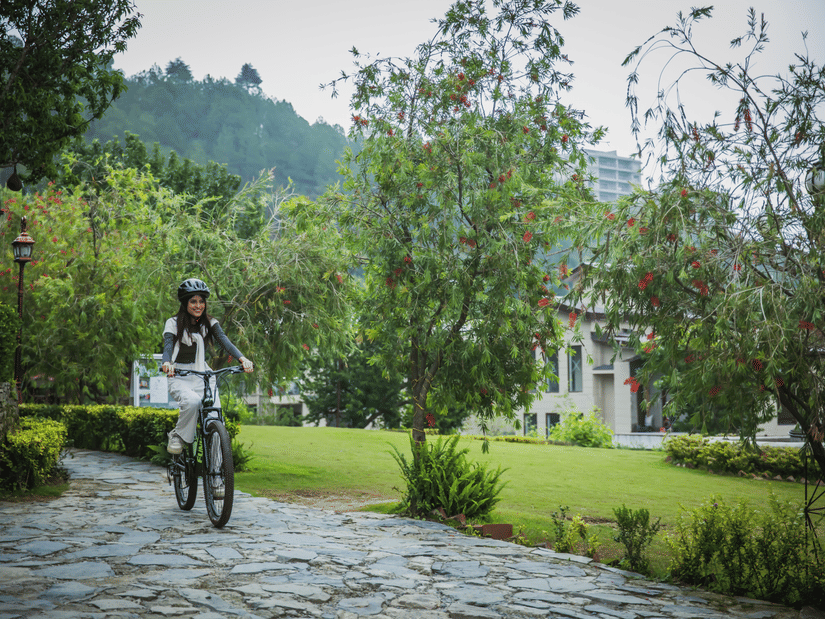 A woman riding a bicycle on stone pathway bordered by lush garden plants, with misty hills in the background, showcasing wellness and recreational activities in the scenic environment of The Manor Sports & Wellness Hotel in Shimla.