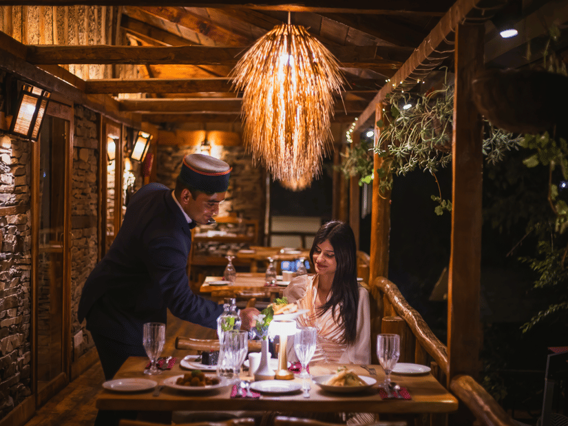 A waiter in a cap serves a woman at a dimly lit, intimate restaurant table with a large, woven light fixture overhead - Manor Sports & Wellness Hotel