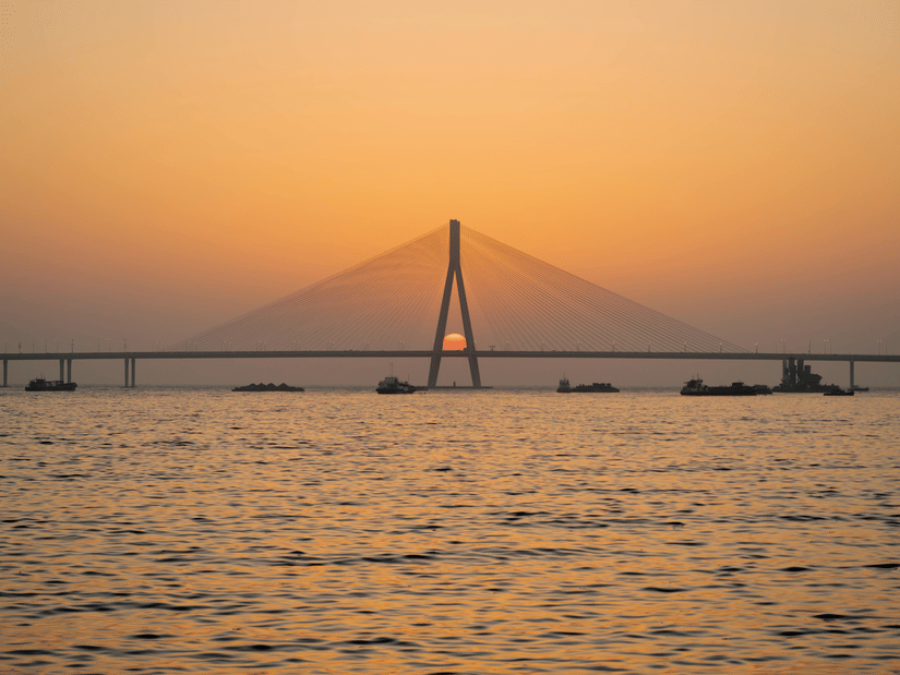 A view from afar of the Bandra-Worli Sea link bridge with the sun setting in the background and the Arabian Sea in the foreground