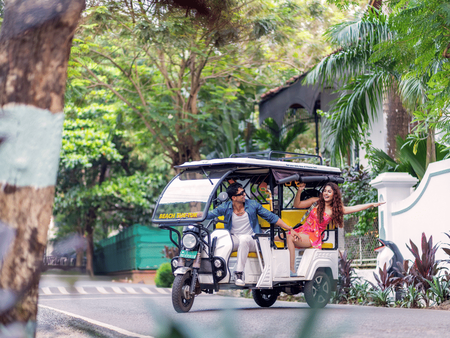 A couple on a colourful tuk-tuk parked under lush trees at a tropical resort entrance | Heritage Village Resorts & Spa, Goa