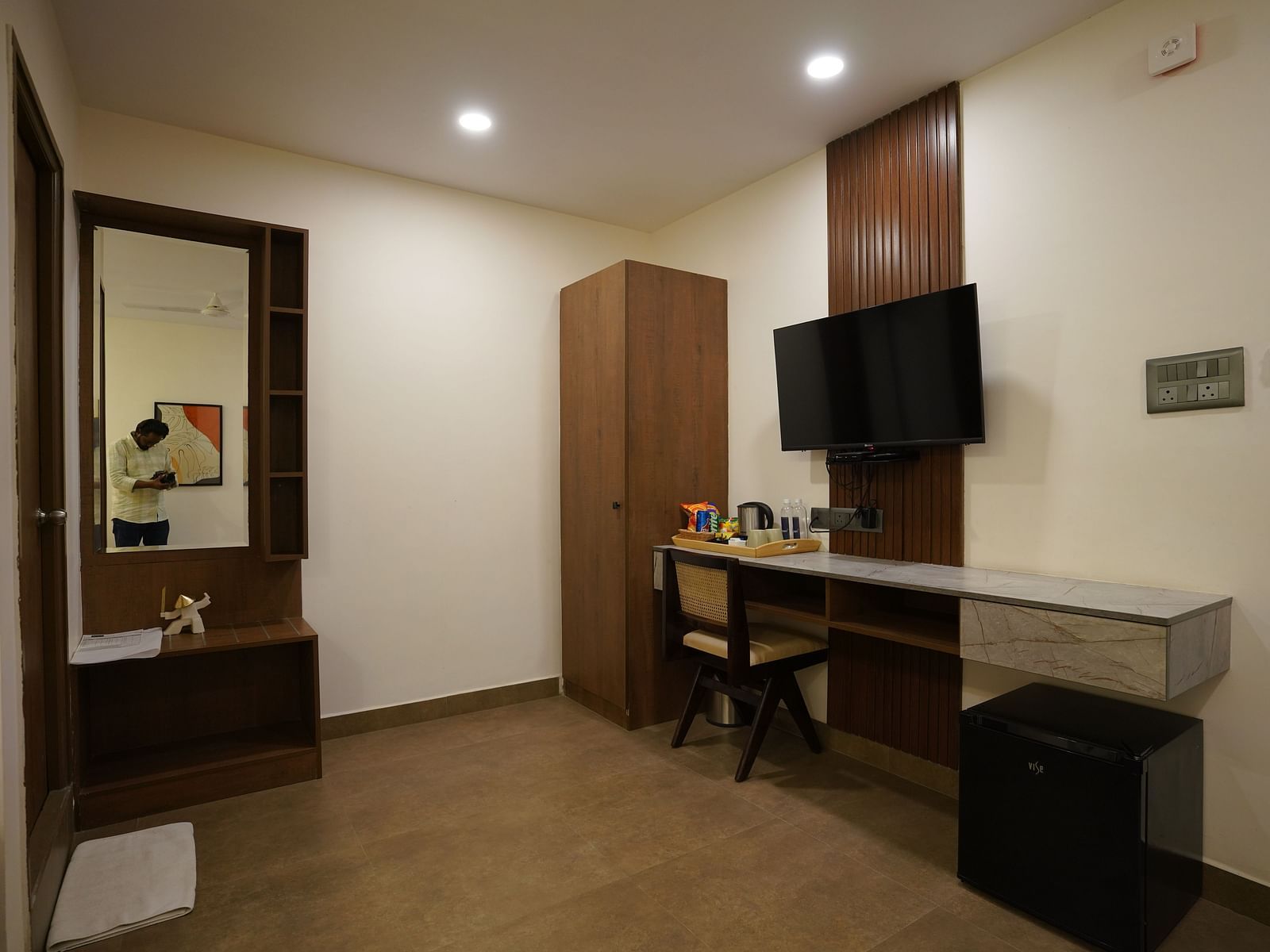 A room with a minimalist design, featuring a wall-mounted TV, a desk with a mirror, and ample lighting at Kyzen Smart Stays.