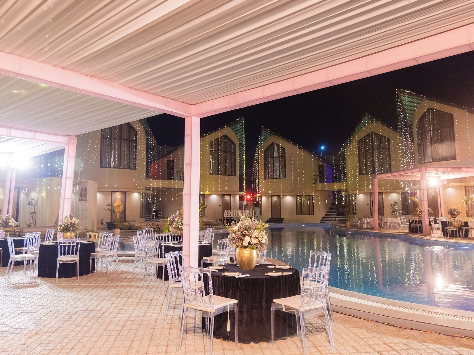 An outdoor wedding reception area is set up next to a pool at night with tables and chairs under a canopy | The Qualia Resort Club & Brewery, Udaipur