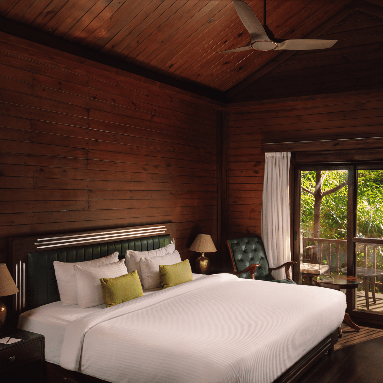 A bedroom at The Ananta Udaipur featuring a king size bed, 2 side tables, armchair, desk, and sliding glass doors leading to a balcony.